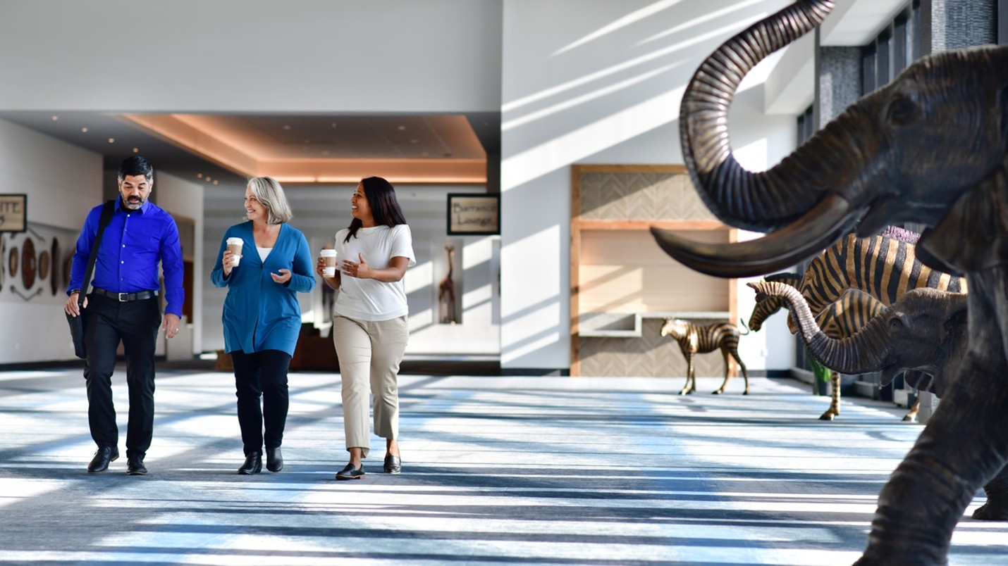 Three people walking down a hallway in a convention center. Two are holding coffee and looking at the third person who is speaking to them.