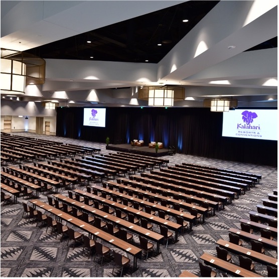 kalahari-mtg-rm A large meeting room set up classroom style with tables and chairs. At the front of the room is a stage with four chairs on it. Two screens hang displaying the Kalahari Resorts and Conventions logo..