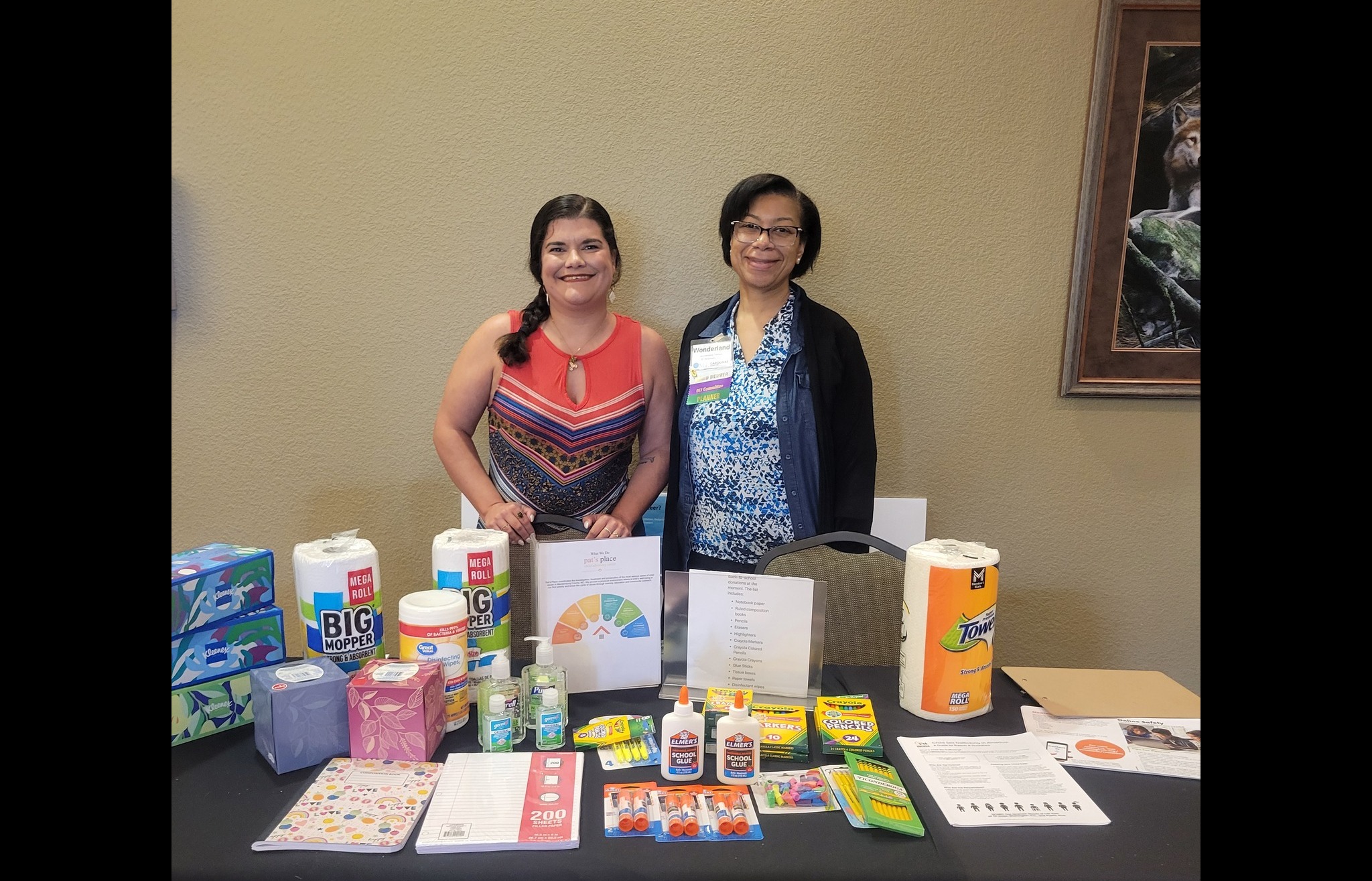 Two people standing together behind a table that has tissues, paper towels, glue, paper, and hand sanitizer on it.