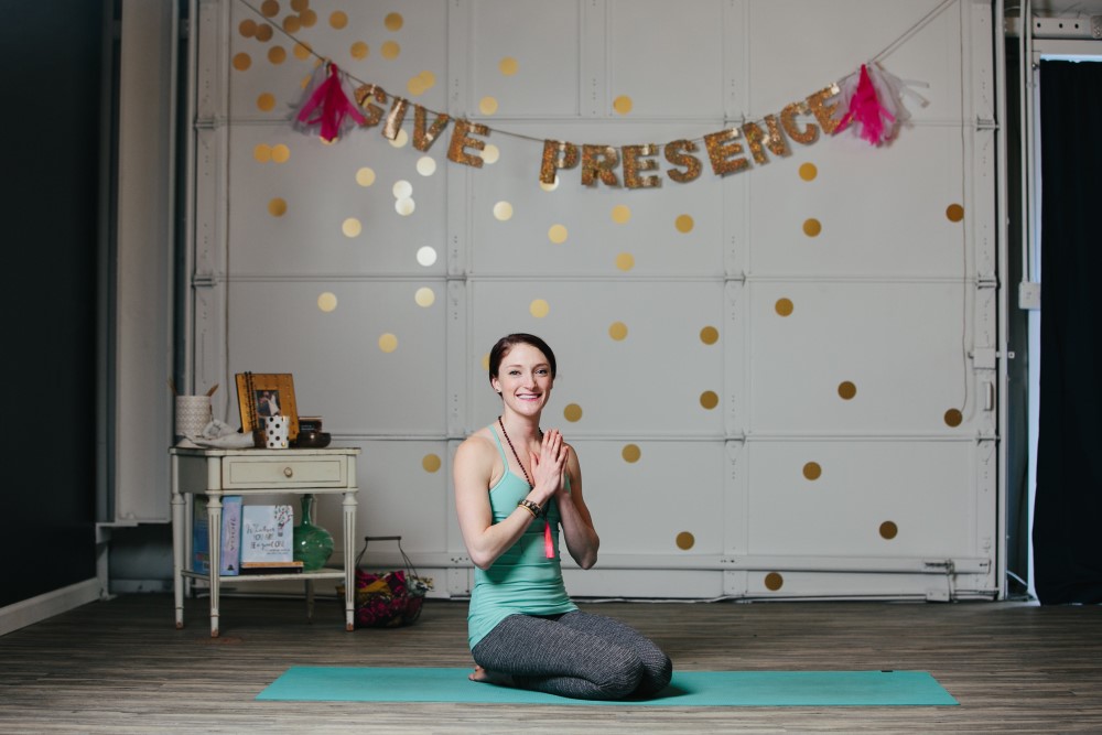 Smiling yoga instructor kneeling on mat in front of wall with banner reading "Give Presence"